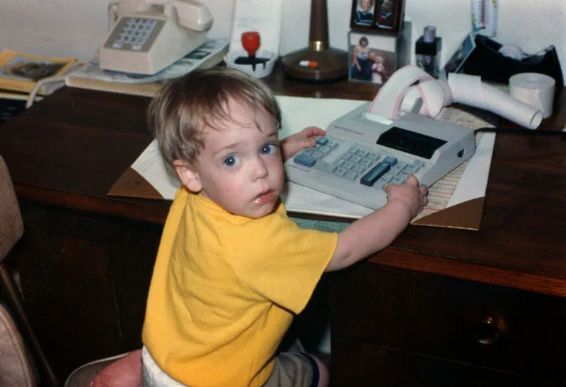 A bar chart illustrates Jared’s early attempts at R programming, showing him as a toddler seated in front of an adding machine. The image conveys the idea that Jared enjoyed data and numbers even in his early years.