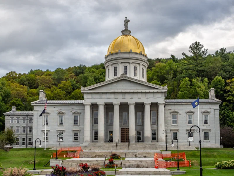 The photograph shows Vermont's state capitol building, a neoclassical structure with a golden dome, set against a backdrop of trees and a cloudy sky. The building represents the real-world data environment that contrasts with the idealized datasets often encountered in academic training.