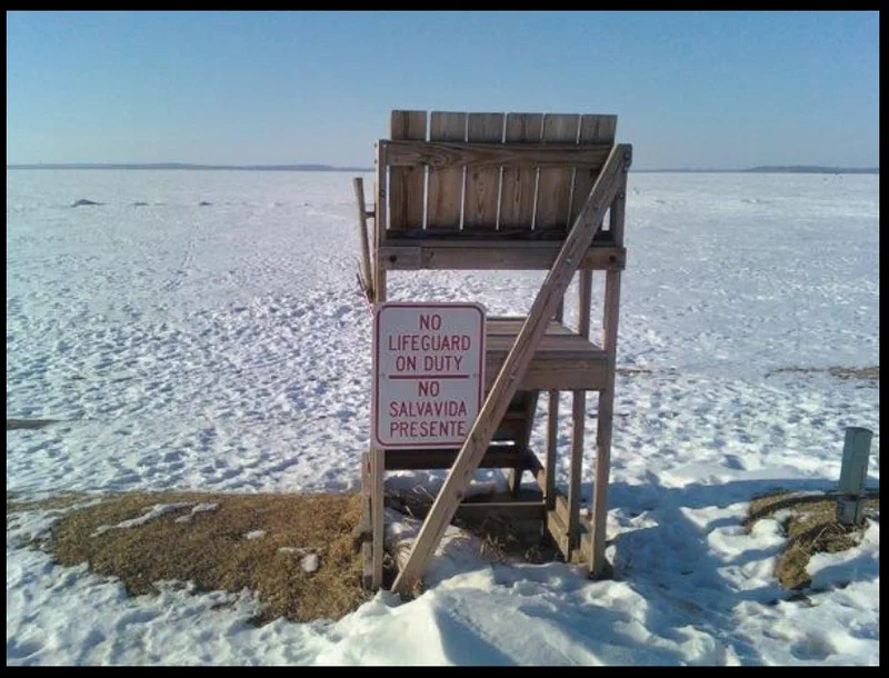 This photograph shows an empty lifeguard stand on a snowy beach in Madison, Wisconsin. The sign reads 'No Lifeguard Duty - No Salvavida Presente,' indicating that swimming is not permitted.